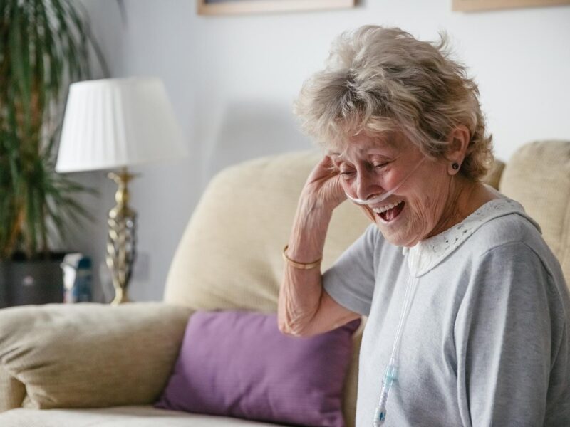A woman with breathing tubes in her nose laughs on a sofa.